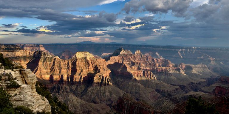 Wide view of the Grand Canyon under a partly cloudy sky, with sunlight highlighting layers of red rock formations and cliffs.