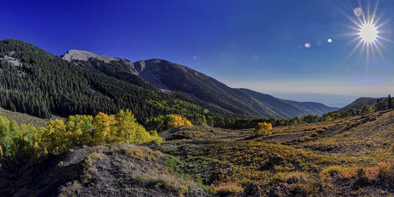 Mountain landscape with dense forests and patches of autumn-colored trees under a clear blue sky, with the sun shining brightly in the upper right corner.