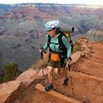 A backpacker in a green shirt with hiking poles treks up the red dirt steps on on a trail in the Grand Canyon