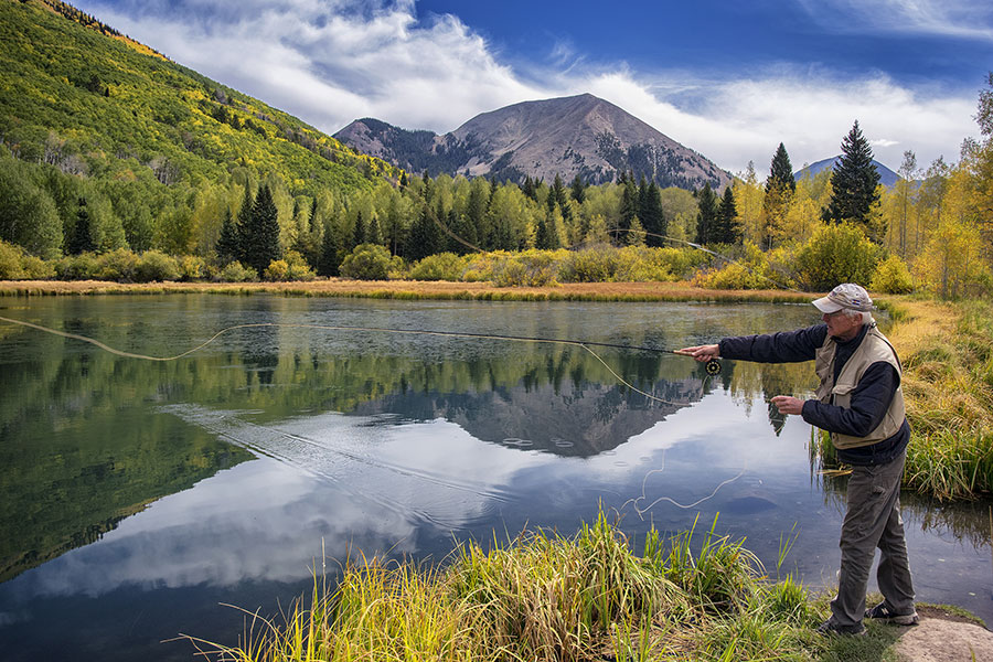 A man casts his fly-rod at the edge of a calm lake, surrounded by trees and mountains under a partly cloudy sky.