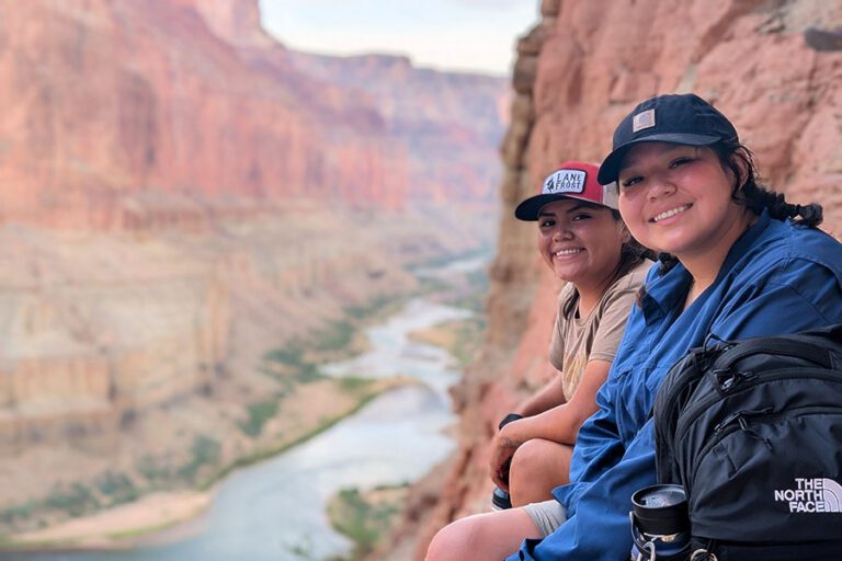 Two people sitting on a rocky ledge overlooking a river winding through a canyon. Both are smiling, wearing caps and casual clothing. One has a camera and backpack beside them.